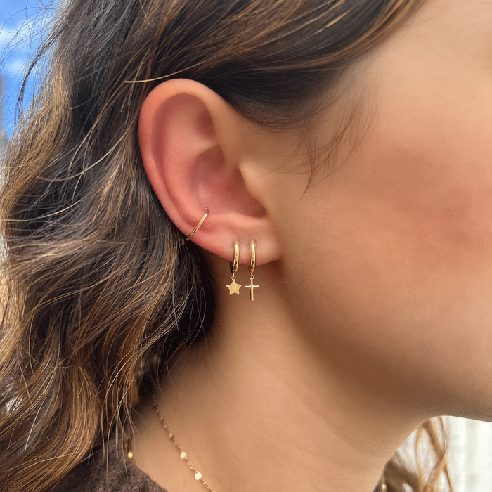 Dainty solid yellow gold ear cuff on model's ear stacked with cross drop huggie and star drop huggie.