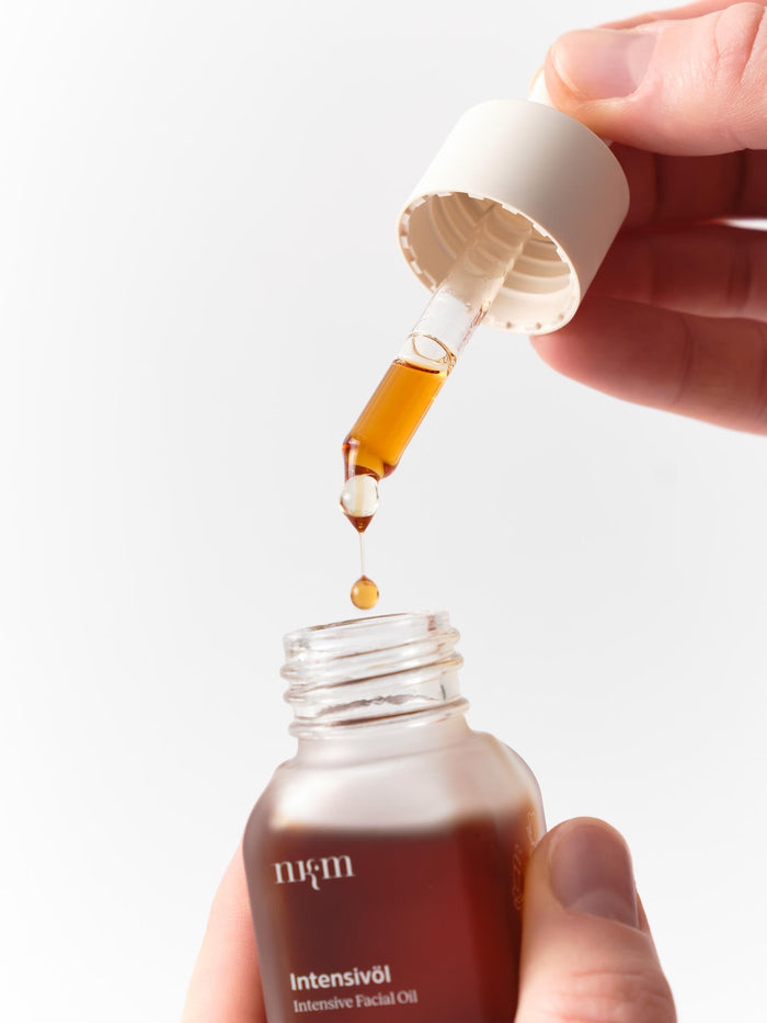 Eine Hand hält eine Pipette über eine geöffnete Flasche mit braunem Intensivöl Neu der nkm Naturkosmetik München GmbH und gibt einen Tropfen zur Vorbeugung vorzeitiger Faltenbildung ab.